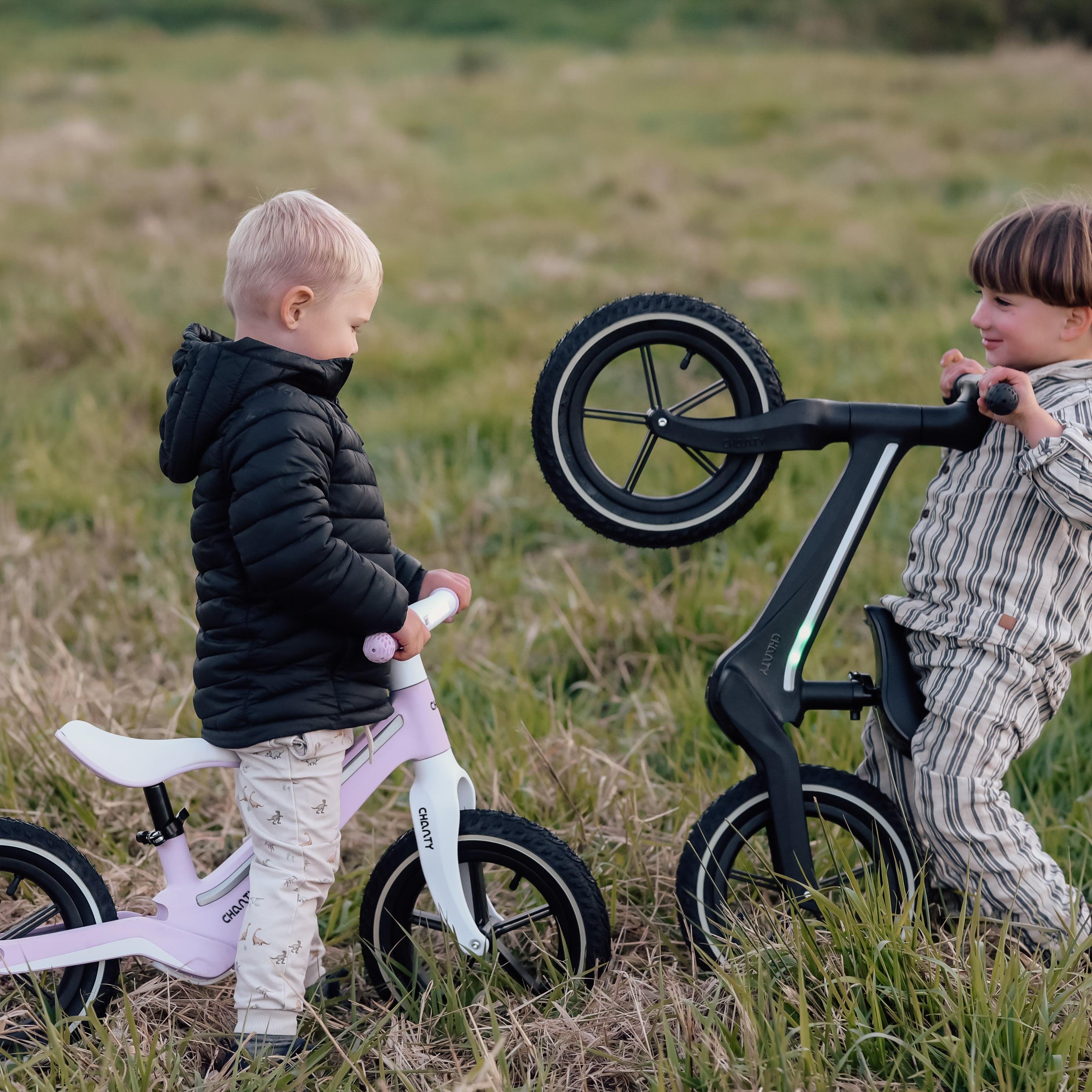 Zwei Jungen spielen auf einer Wiese mit ihren Chanty-Laufrädern, eines in Rosa und eines in Schwarz.