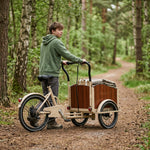 Eine Person schiebt ein beiges Lastendreirad mit Holzkiste auf einem Waldweg.
